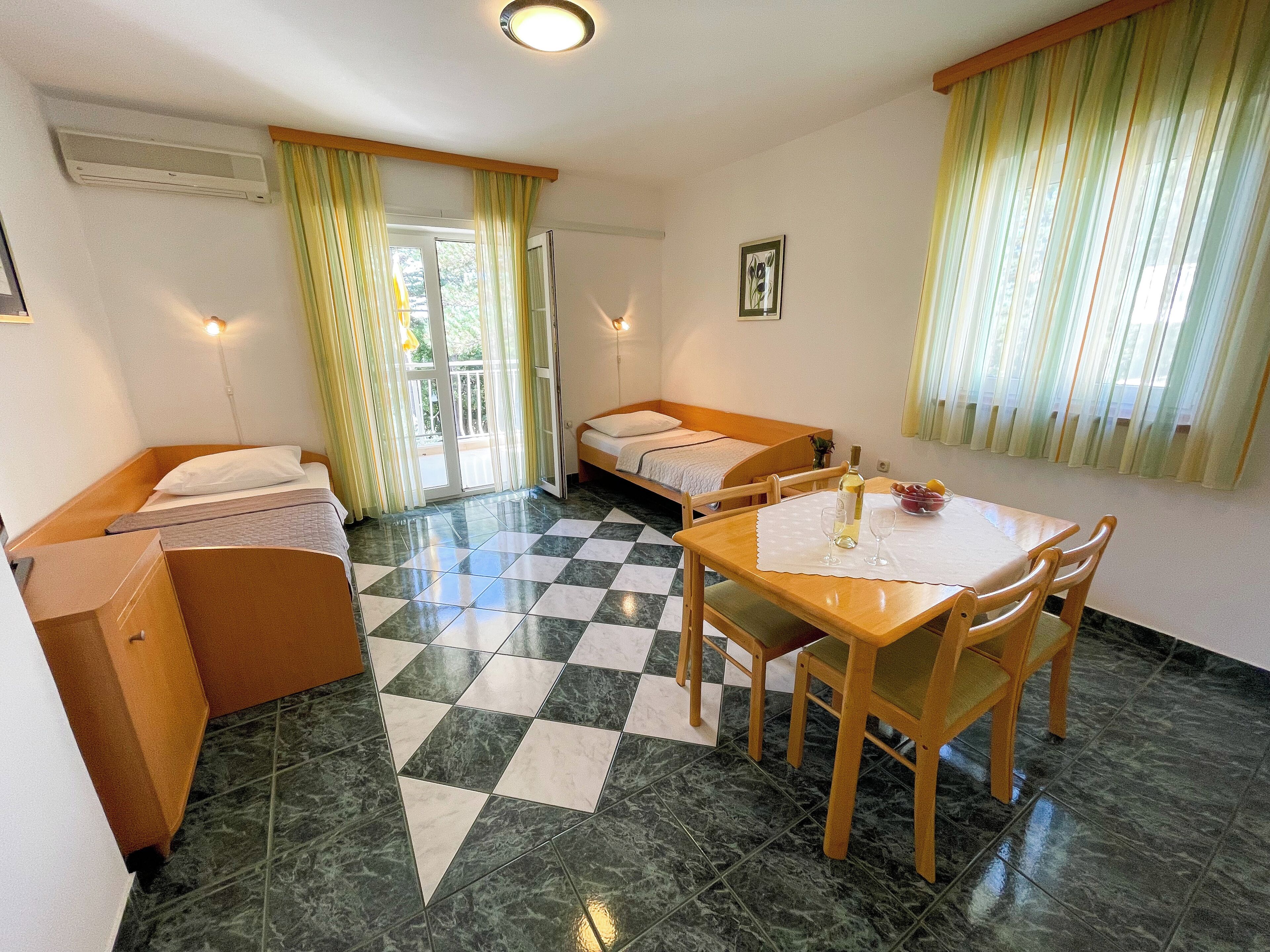 Una habitación con piso a cuadros, dos camas individuales, una mesa de madera con sillas y una ventana con cortinas.