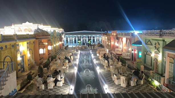 Ein gut beleuchteter Essbereich im Freien mit Tischen und Stühlen, einer zentralen Wasserspiele und ein Gebäude mit aufwendiger Architektur im Hintergrund.