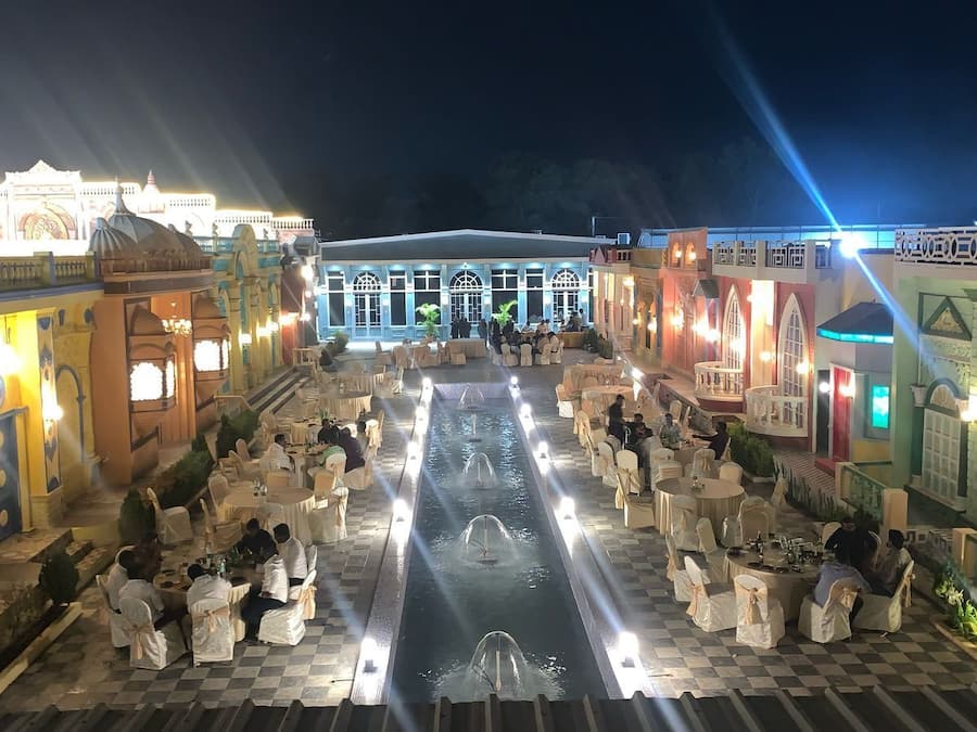 A well-lit outdoor dining area with tables and chairs, a central water feature, and a building with ornate architecture in the background.
