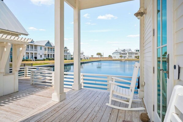 A wooden deck with a white rocking chair, a pool, and a view of multi-story buildings in the background.