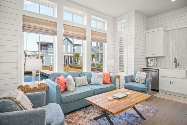 A spacious living room with a blue sofa set, a wooden coffee table, and a view of a pool and neighboring houses. - Charming Lakefront Cottage with Breathtaking Views in Cinnamon Shore South (Port Aransas)