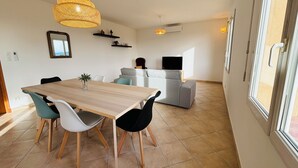 Een moderne eetruimte met een houten tafel en stoelen, een grijze bank en een televisie op de achtergrond.