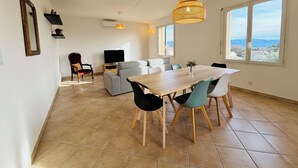 Een ruime woonkamer met een eettafel, stoelen, een bank en een televisie. Grote ramen bieden uitzicht op het omliggende landschap.
