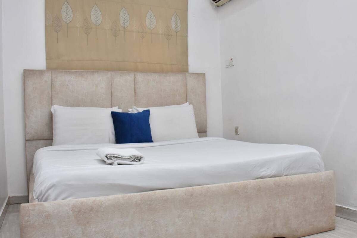 Una cama bien hecha con sábanas blancas y una almohada azul, un cabecero color beige y una pared con un estampado de hojas.