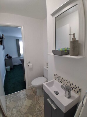 A bathroom with a toilet, sink, and shower area. There is a mirror above the sink and a shelf with toiletries. The floor has patterned tiles.