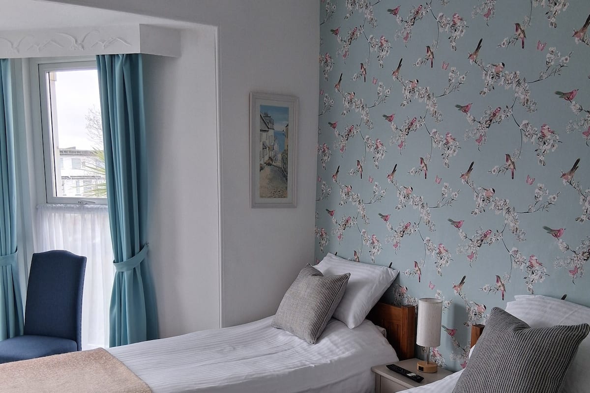 A hotel room with two beds, a chair, a nightstand with a lamp, and a floral wallpaper.