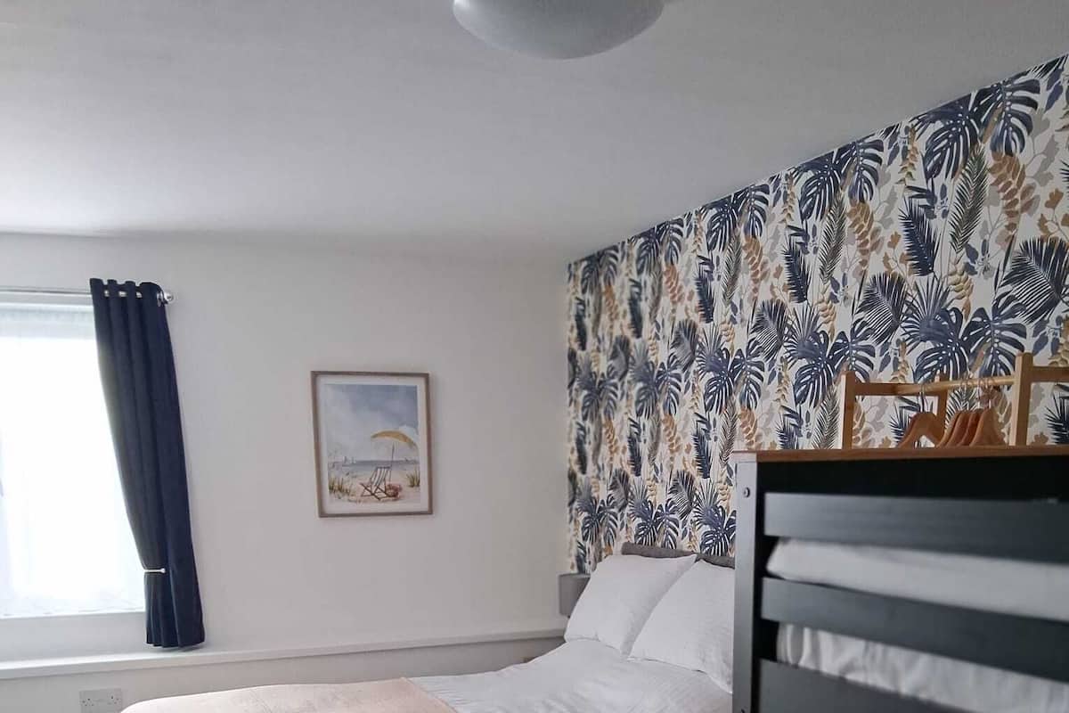 A bedroom with a bunk bed, a single bed, a patterned wall, a window with curtains, and a framed picture on the wall.