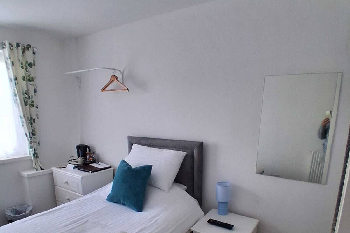 A neatly made bed with white linens and a blue pillow, a bedside table with a remote control, a small nightstand with a coffee maker, and a mirror on the wall.