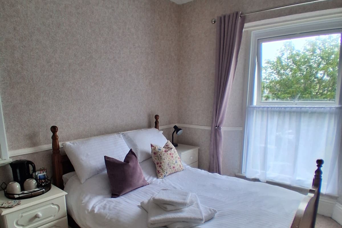 A neatly made bed with pillows, a bedside table with a lamp, and a window with curtains.