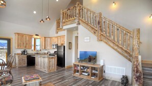 Una cocina amplia con armarios de madera, una isla central y una estufa integrada. Una escalera de madera con un diseño rústico conduce a una sala de estar con televisor y estantería.