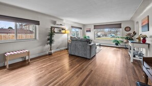 A spacious living room with hardwood floors, a grey sofa, a wooden bench, and large windows offering a view of the outside. - Hot Tub & Gym | Minutes to Red Rocks Ampitheater | Sauna (Lakewood)