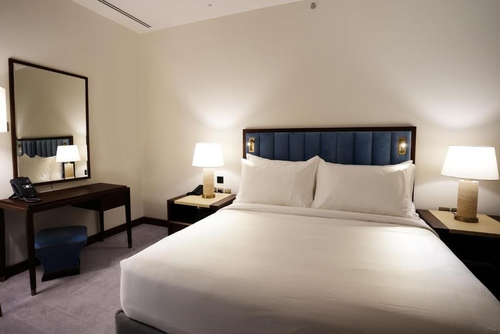 A hotel room with a large bed, a desk, a chair, two bedside tables with lamps, and a large mirror on the wall.