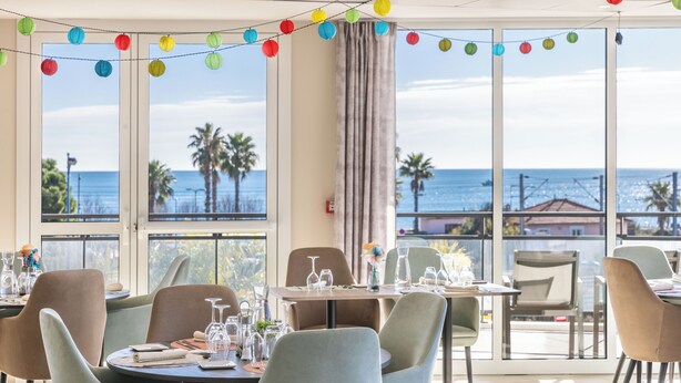 Un espace repas avec des tables préparées pour deux personnes, de grandes fenêtres offrant une vue sur la mer et des lanternes en papier colorées suspendues au plafond.