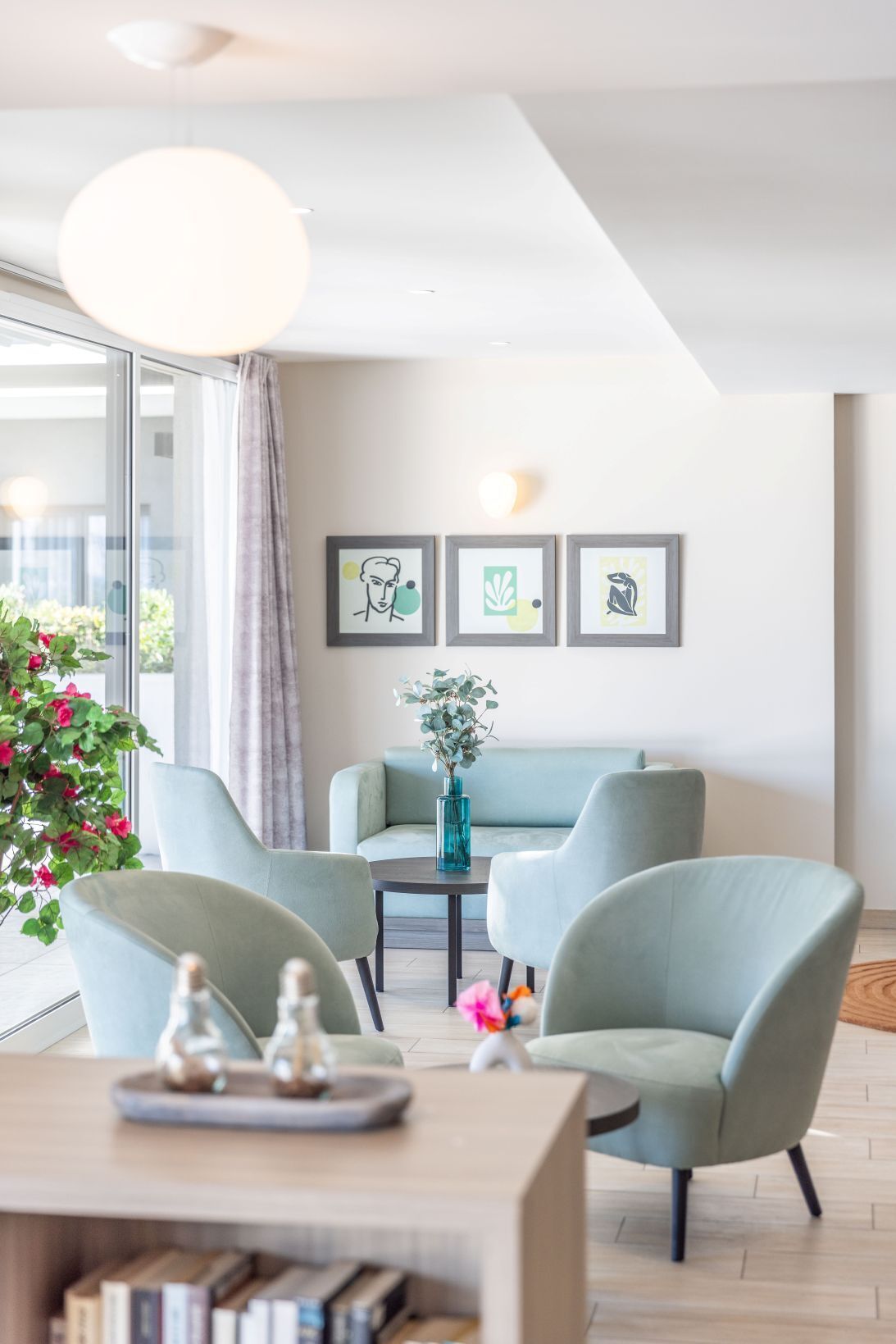 Un salon moderne comprenant un canapé, deux fauteuils, une table basse et un vase avec des fleurs. Trois œuvres d’art encadrées ornent le mur, et une grande fenêtre est agrémentée de rideaux.