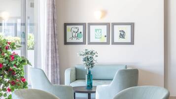 Un salon moderne comprenant un canapé, deux fauteuils, une table basse et un vase avec des fleurs. Trois œuvres d’art encadrées ornent le mur, et une grande fenêtre est agrémentée de rideaux.