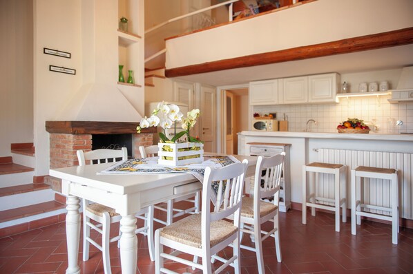A dining area with a white table and chairs, a fireplace, and a kitchen in the background.
