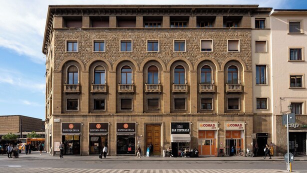 Un immeuble à plusieurs étages, doté d’une façade en pierre et abritant divers commerces, notamment un bar et des restaurants.