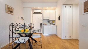 Une cuisine compacte avec des meubles blancs, un coin repas doté d’une table en verre et de chaises en métal, et un plancher en bois.