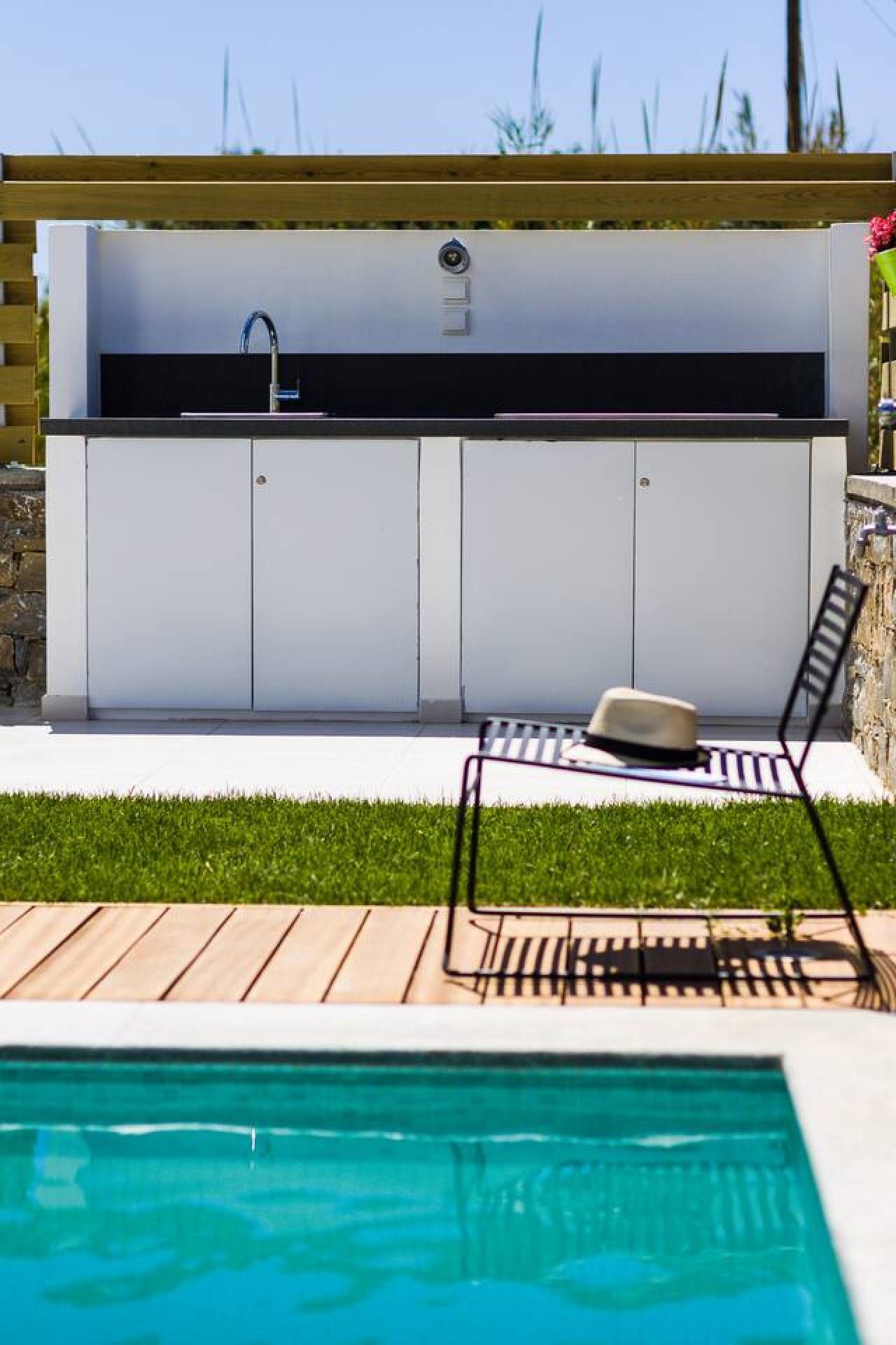 A modern outdoor kitchen with a built-in sink and cabinets, a poolside lounge area with a chair and a hat, and a clear view of the pool.