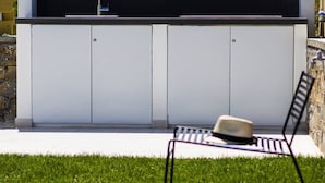 Una cocina moderna al aire libre con pileta y muebles bajo techo, un área de descanso junto a la piscina con una silla y un sombrero, y una vista despejada de la piscina.