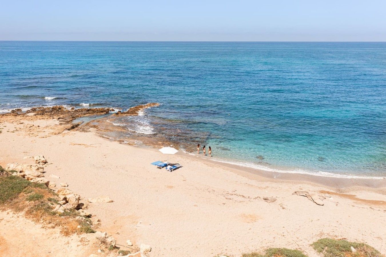 Une plage à l’eau d’un bleu limpide, avec un rivage sablonneux, où quelques personnes se promènent.