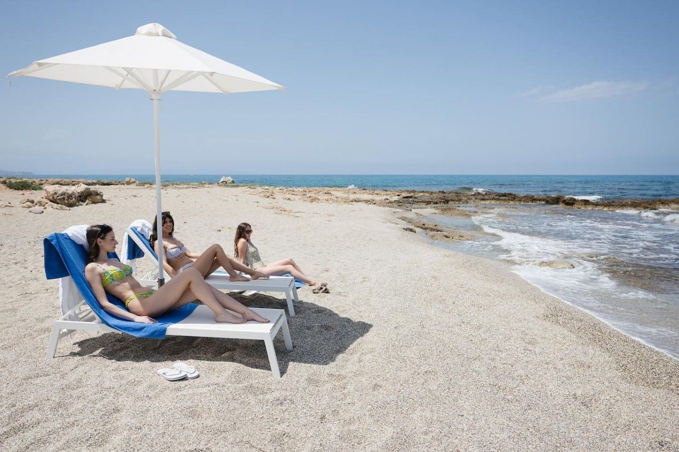 En strandscene med tre liggestole under en hvid parasol, en stenstrand og havet i baggrunden.