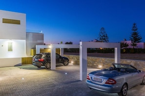 Un edificio moderno con una zona de estacionamiento techada, una camioneta SUV negra y un automóvil descapotable plateado.