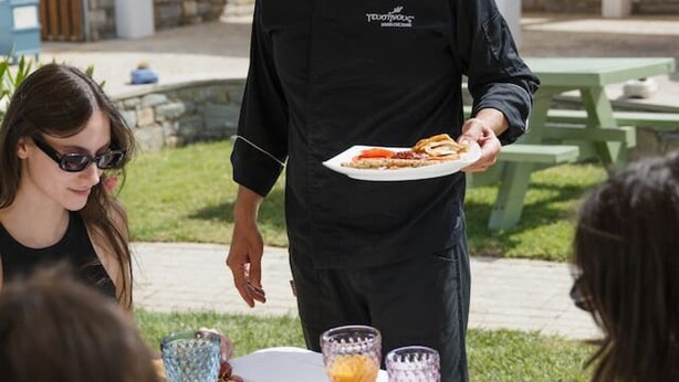 A chef serving food to guests at an outdoor table with a white tablecloth, surrounded by greenery and a building in the background. - Persi by the beach! Thalasses villas (Rethymno)