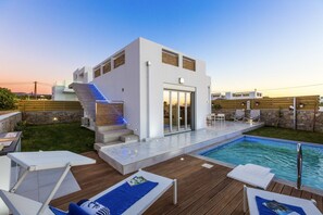 A modern outdoor pool area with a wooden deck, lounge chairs, and a staircase leading to a white building with large windows. - Persi by the beach! Thalasses villas (Rethymno)