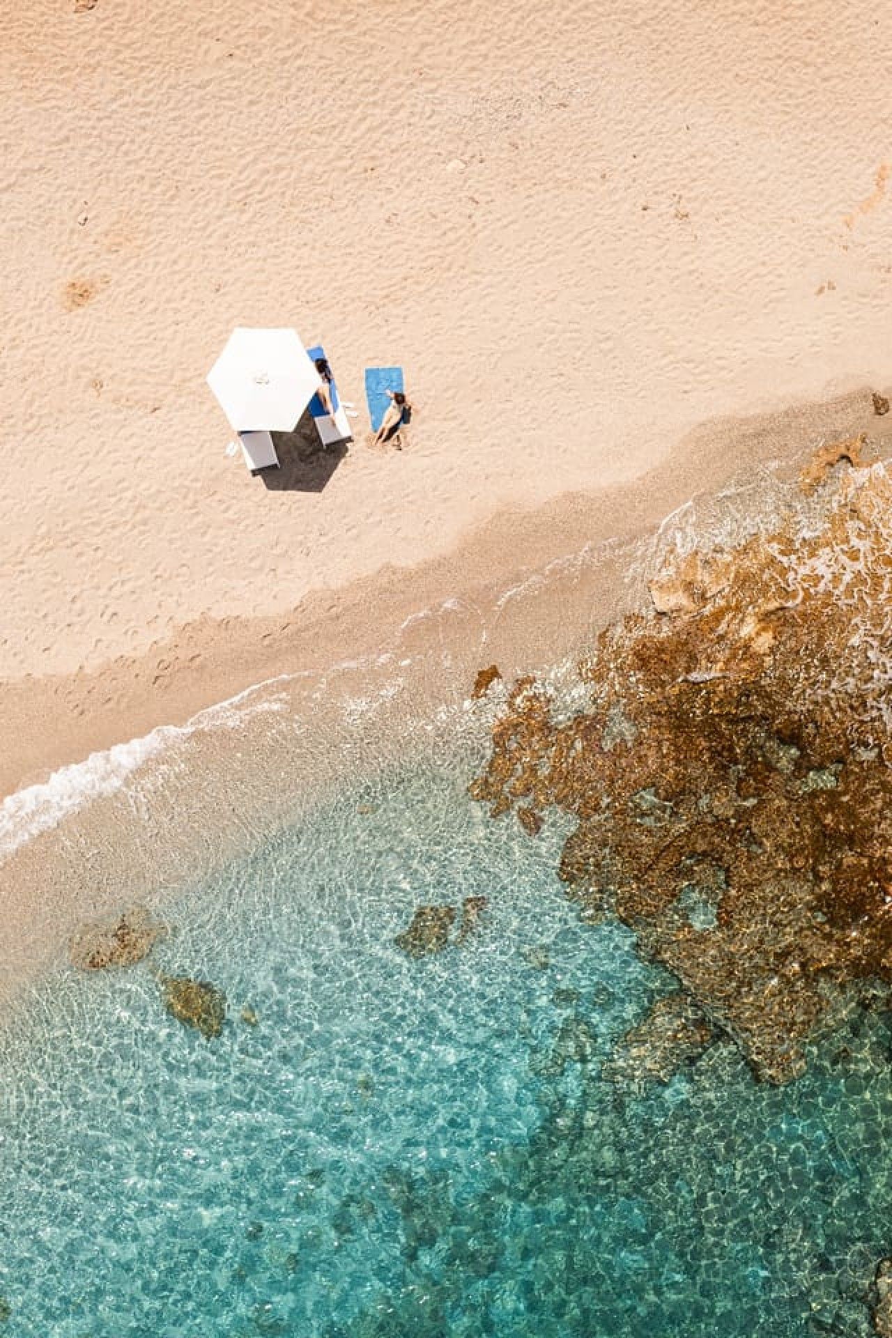 An aerial view of a beach with clear water, a person with a white umbrella, and a blue beach chair.