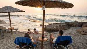 A beach scene with two people seated under a thatched umbrella, a table with a bottle and glasses, and a rocky shoreline. - Persi by the beach! Thalasses villas (Rethymno)