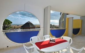 A room with a round table and chairs, a red tablecloth, and a view of a beach with a thatched-roof structure.