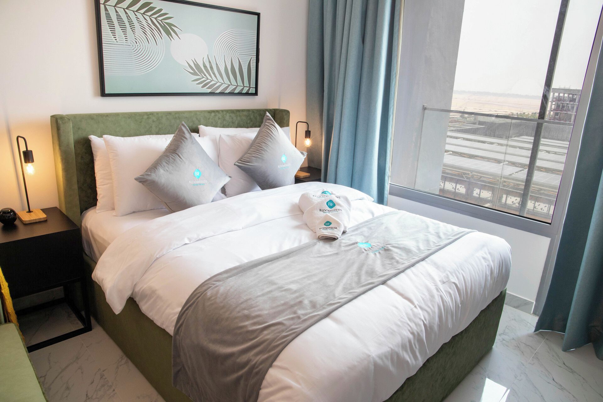 A hotel room with a bed, pillows, a nightstand, a lamp, a painting, and a view of the city.