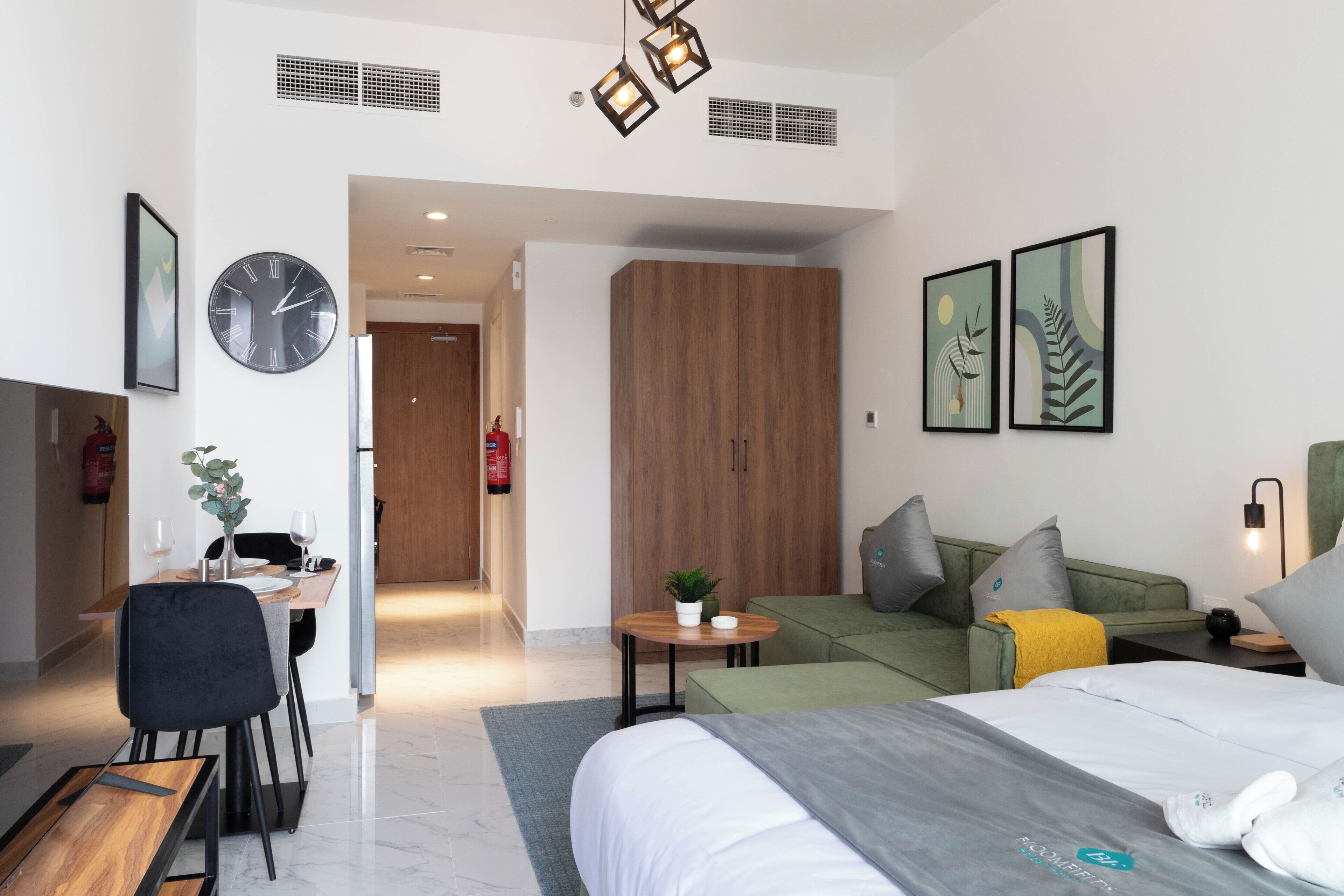A modern hotel room with a bed, a sofa, a dining table with chairs, a clock, and artwork on the walls.