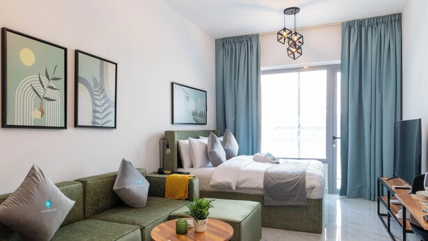 A modern hotel room with a green sofa, a bed with white bedding, a wooden side table, and a large window with curtains. - Bloomfields Euphoric STU in Oasis Masdar (Abu Dhabi)