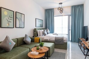 Une chambre d’hôtel moderne avec un canapé vert, un lit avec une literie blanche, une table de chevet en bois et une grande fenêtre avec des rideaux.