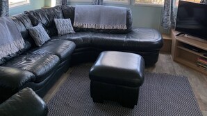 A modern living room with a black leather sectional sofa, a black ottoman, and a television on a wooden stand. Large windows with patterned curtains allow natural light and views of the outdoors.