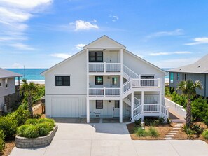 A two-story beachfront house with a balcony, a front porch, and a driveway leading to the entrance.