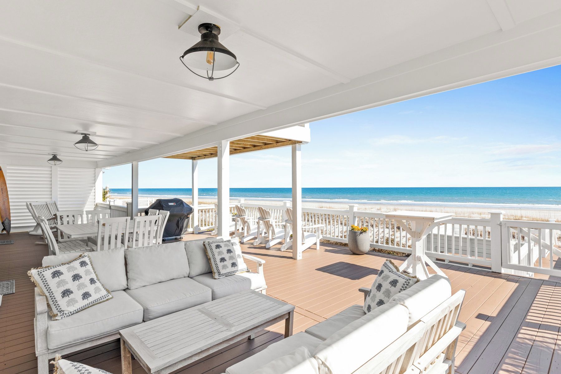 A covered outdoor seating area with white furniture, a grill, and a view of the beach.