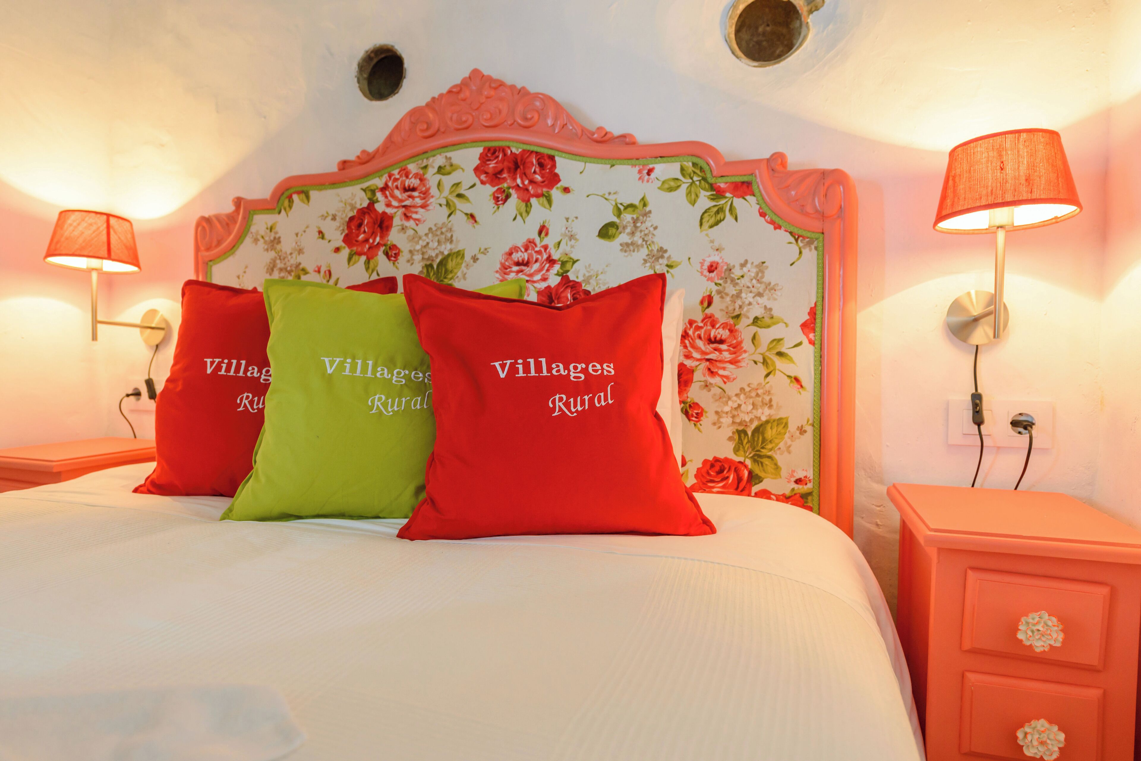 A bed with floral headboard, red and green pillows with "Villages Rural" text, and two bedside lamps.