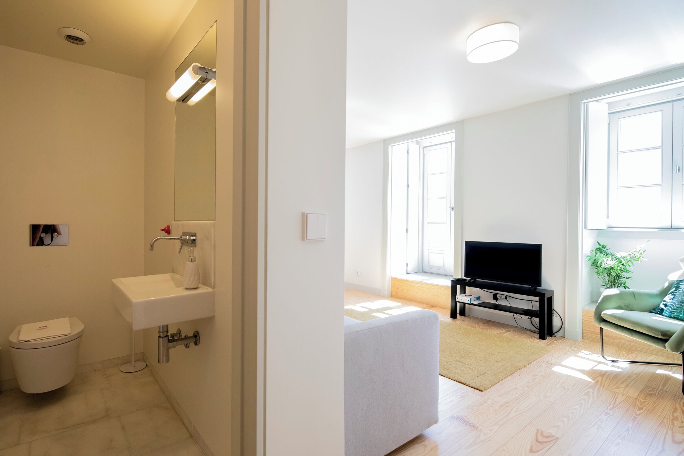 Un baño moderno con inodoro, lavabo y espejo. La habitación tiene un ventanal grande y un televisor de pantalla plana sobre un mueble.