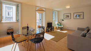 Una sala de estar moderna con una mesa de comedor de vidrio, un sofá gris y un televisor. Hay dos cuadros en la pared y una ventana con vistas a edificios.