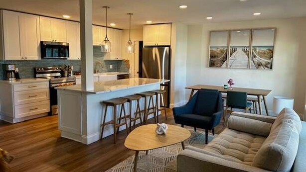 A modern kitchen with white cabinets, a central island with bar stools, a dining area with a table and chairs, and a living room with a sofa and a coffee table. - NEW Stunning Home-close to Beaches-pet Friendly (Largo)