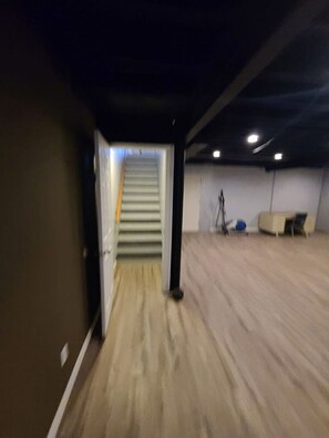 Ein geräumiges Zimmer mit Holzboden, einer Treppe zu einer oberen Ebene und einer Decke mit eingelassenen Leuchten.