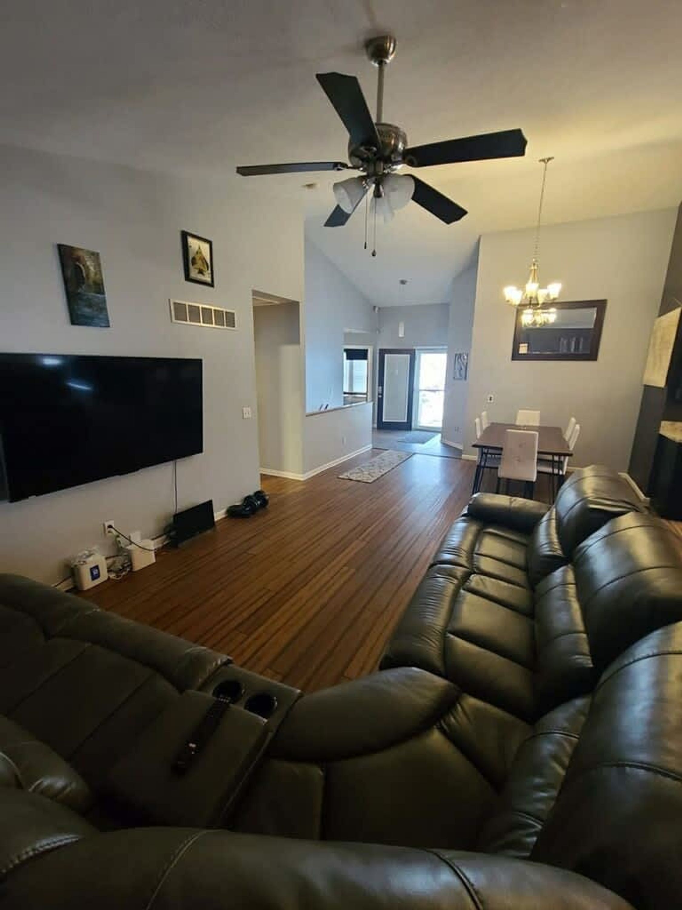 A living room with a large flat-screen TV, a ceiling fan, a dining area with a table and chairs, and a hallway leading to a door.