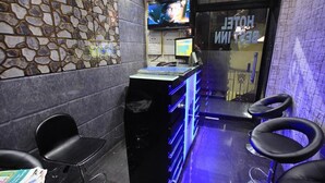 Zona de recepción de un hotel con pared de piedra, mostrador de recepción con computadora y televisor, y tres taburetes negros.
