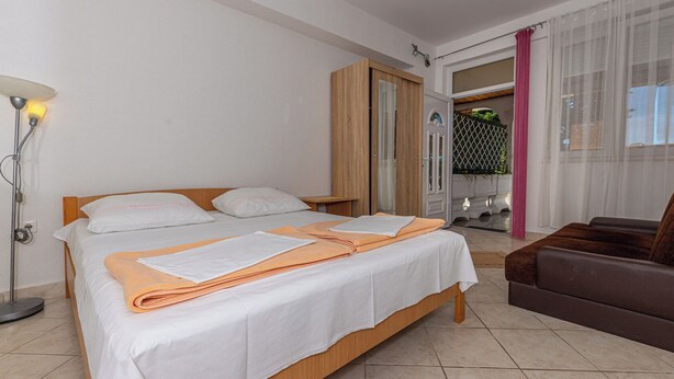 Een hotelkamer met een tweepersoonsbed, een houten garderobe, een tafeltje, een vloerlamp en uitzicht buiten door een raam met dunne gordijnen.