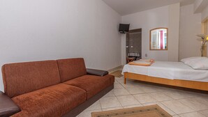 Een slaapkamer met een bruine bank, een bed met witte lakens, een tafeltje en een televisie aan de muur.