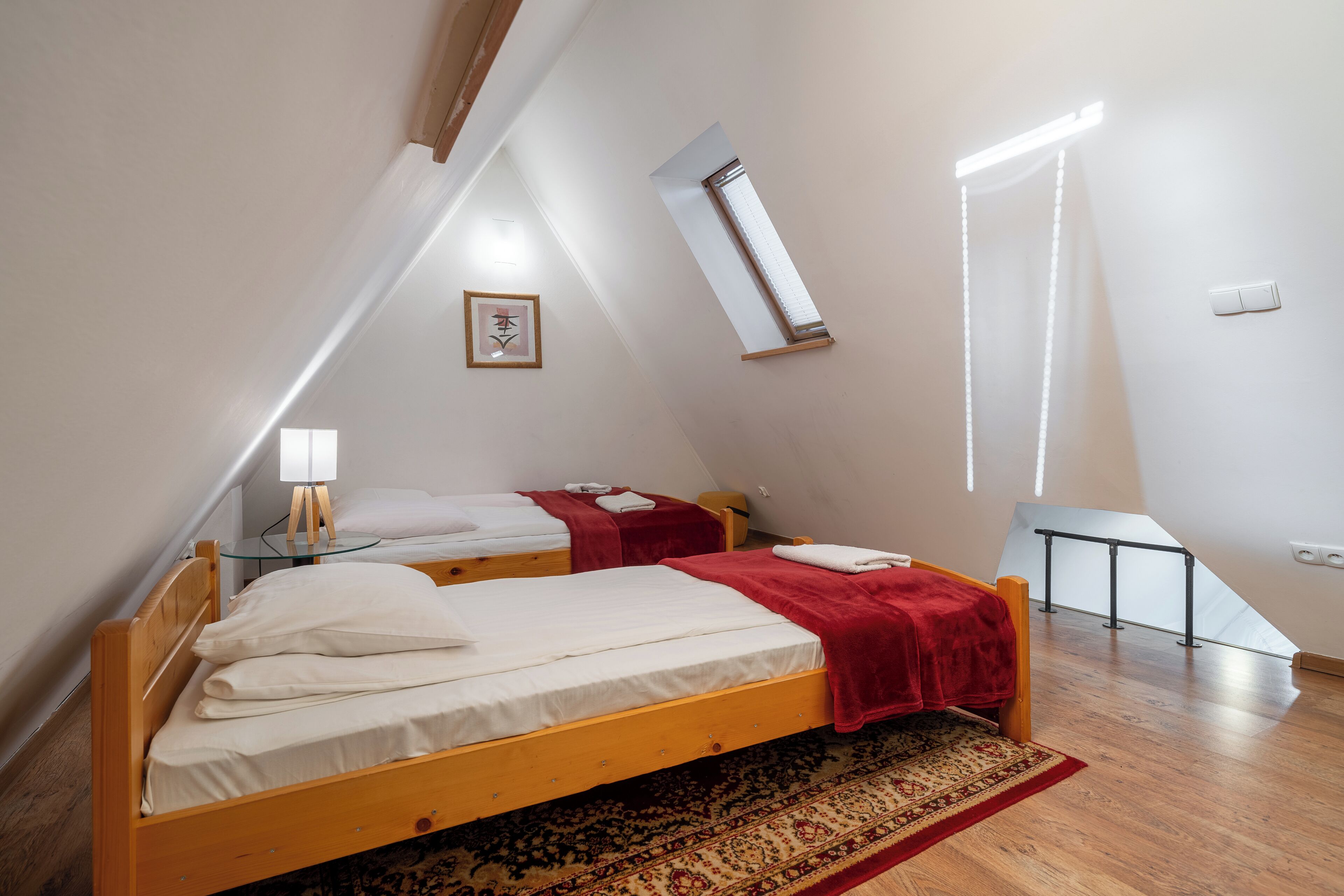 A small, triangular room with two wooden beds, a red blanket, a small table, and a patterned rug.