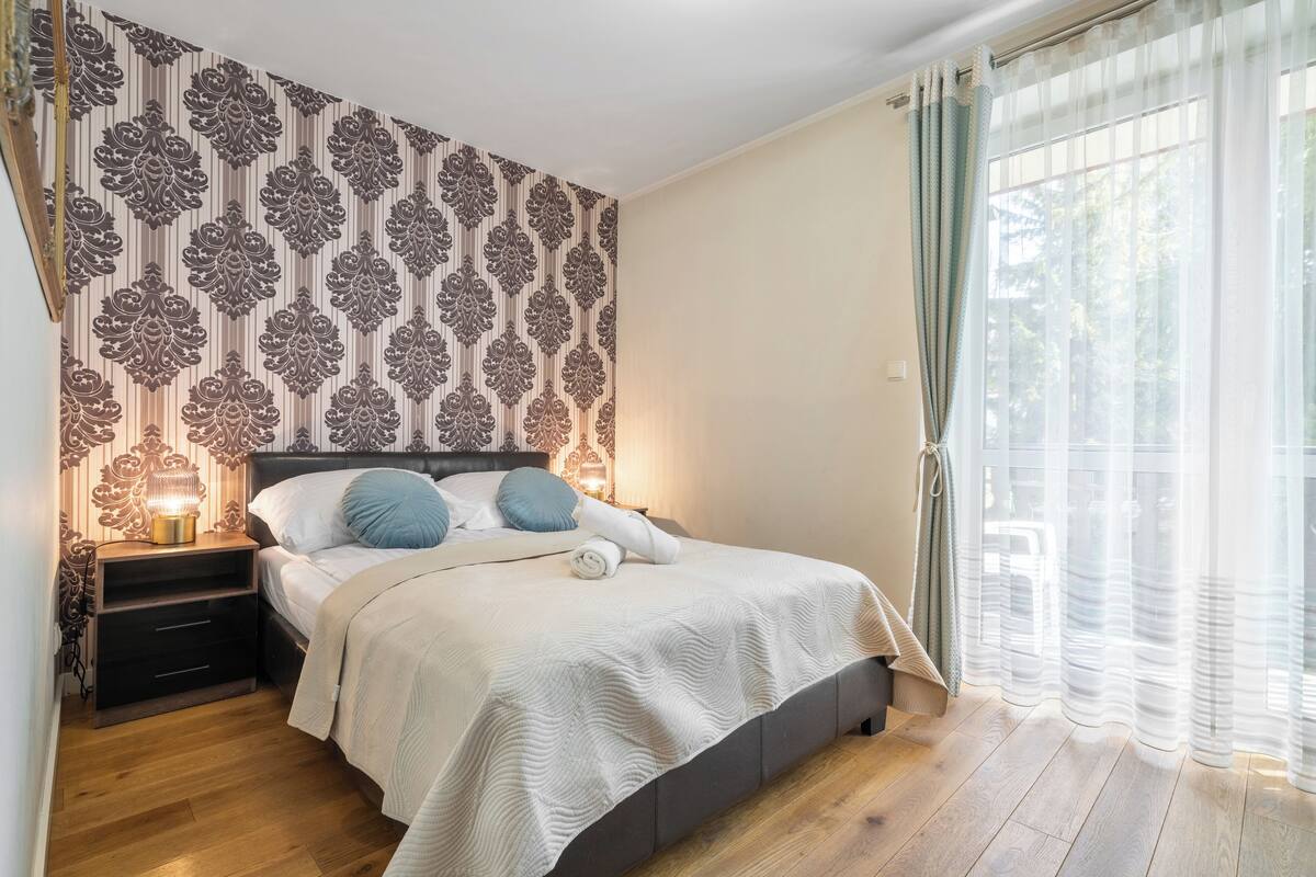 A bedroom with a patterned wallpaper, a bed with pillows, a nightstand with a lamp, and a window with curtains.